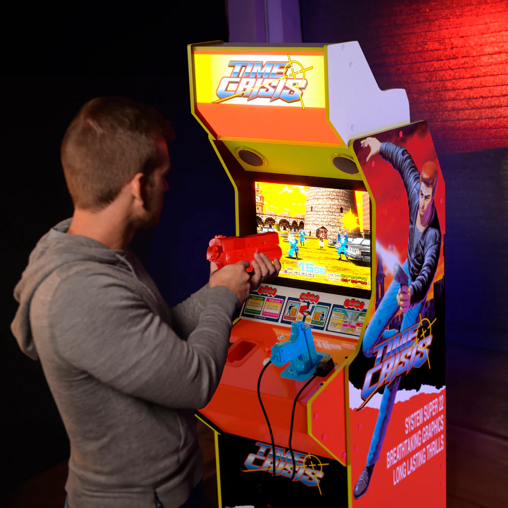 Férfi Time Crisis arcade játékkal játszik. Piros pisztolyt tart, a játék képernyője látható. A játékgép narancssárga és fehér. Poszter az oldalon.