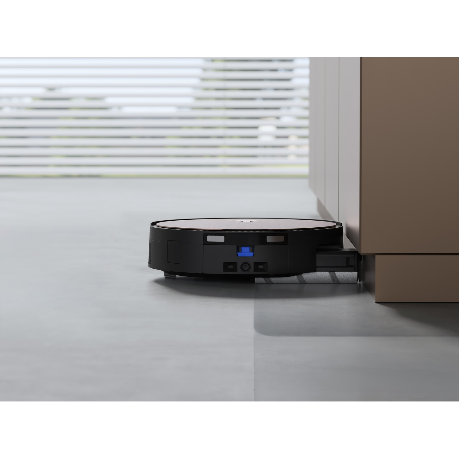 Un aspirapolvere robot nero e argento è posizionato vicino a un armadio beige. Lo sfondo mostra persiane e una finestra.