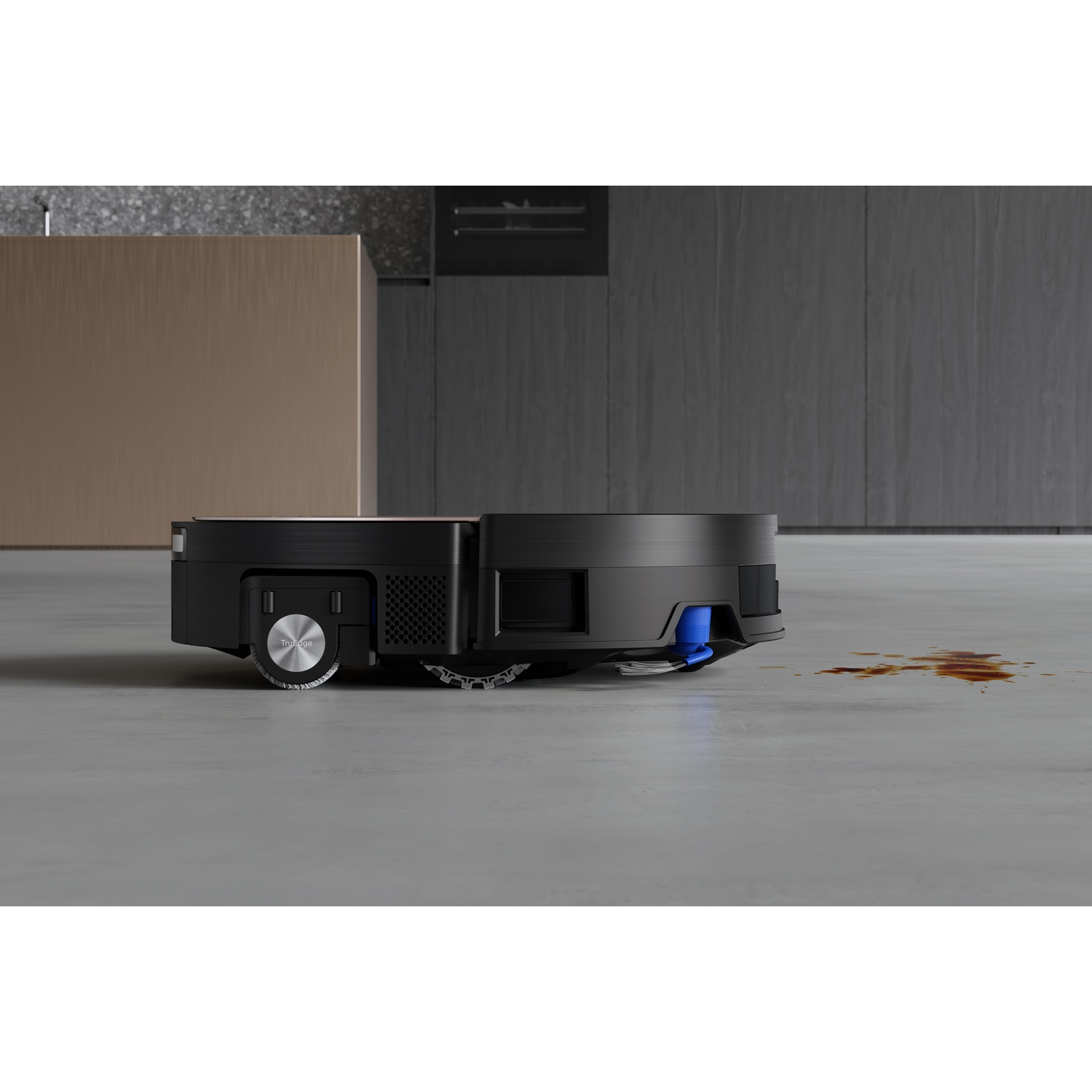 Un aspirapolvere robot nero su un pavimento in cemento. Macchia marrone visibile. Lo sfondo mostra mobili da cucina e un pannello di legno.