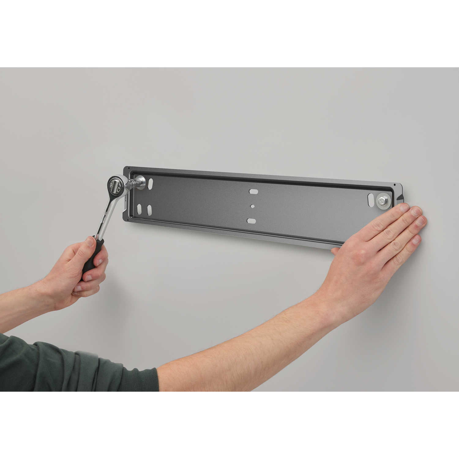 Handen houden een moersleutel en een metalen beugel op een grijze muur. Eén hand draait een bout vast, de andere houdt de beugel vast.