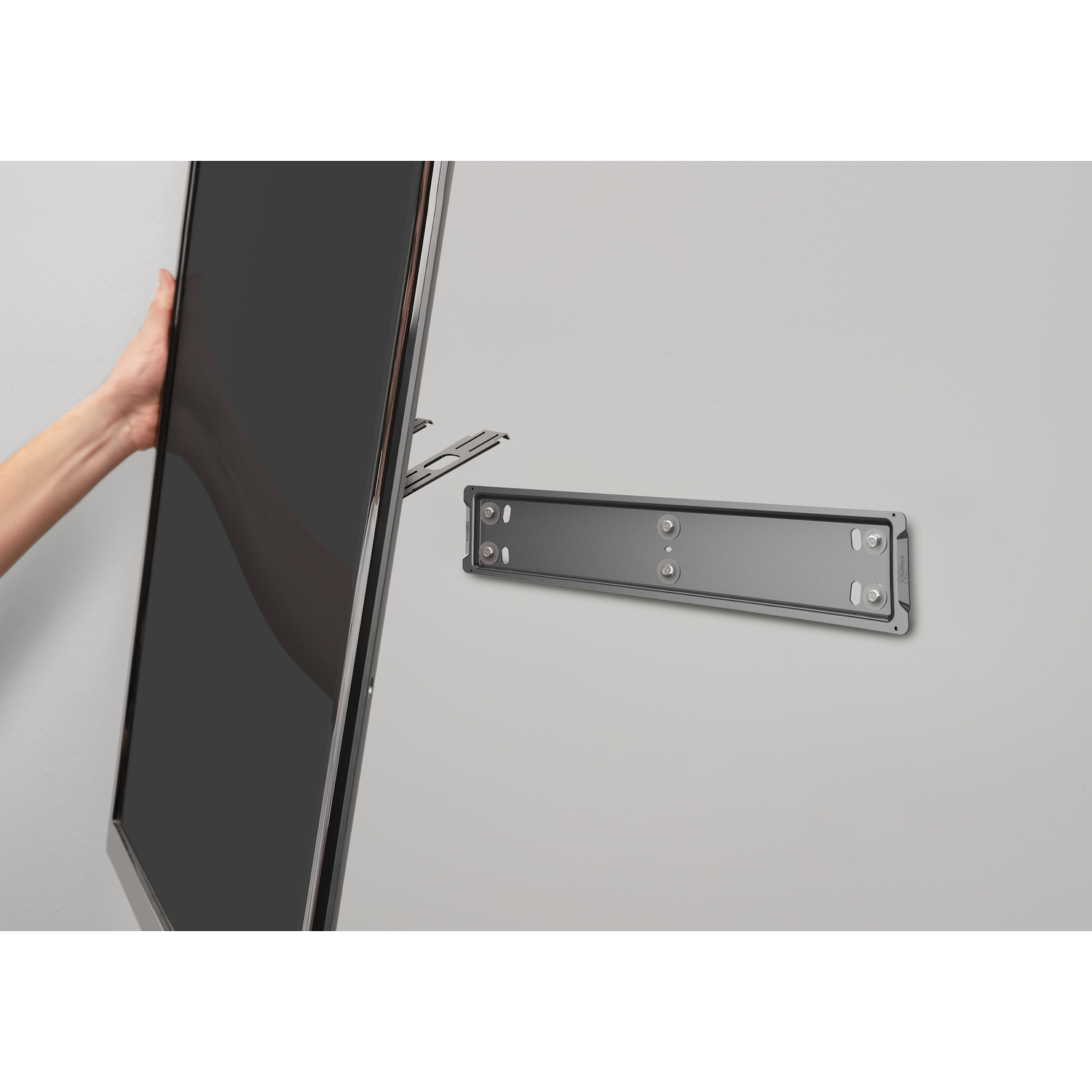 Een hand houdt een zwart tv-scherm vast, in de buurt van een muurbeugel. De muur is lichtgrijs.