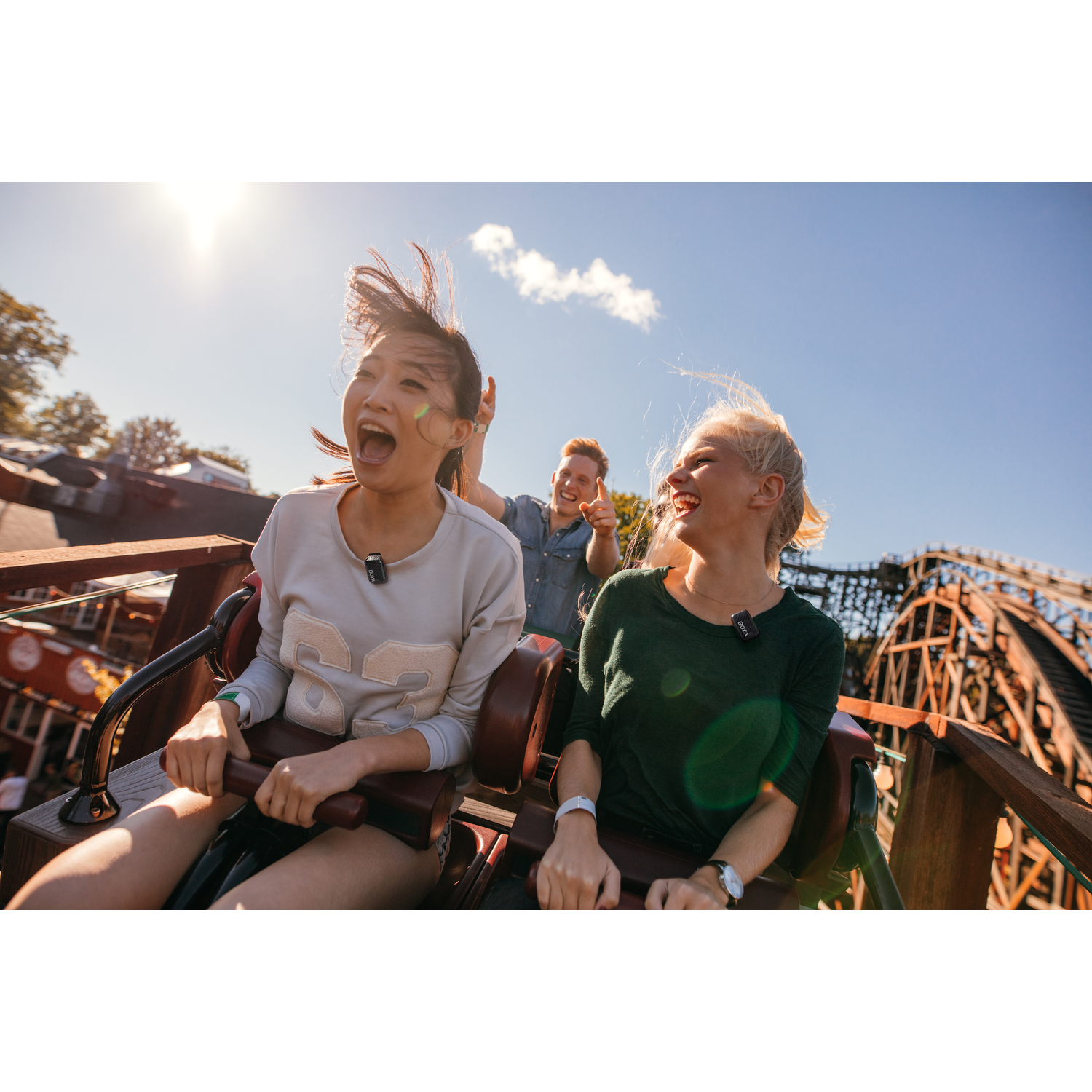 Three people on a rollercoaster. Two women scream, wind blowing their hair. A man behind them points. Wooden track in the background.