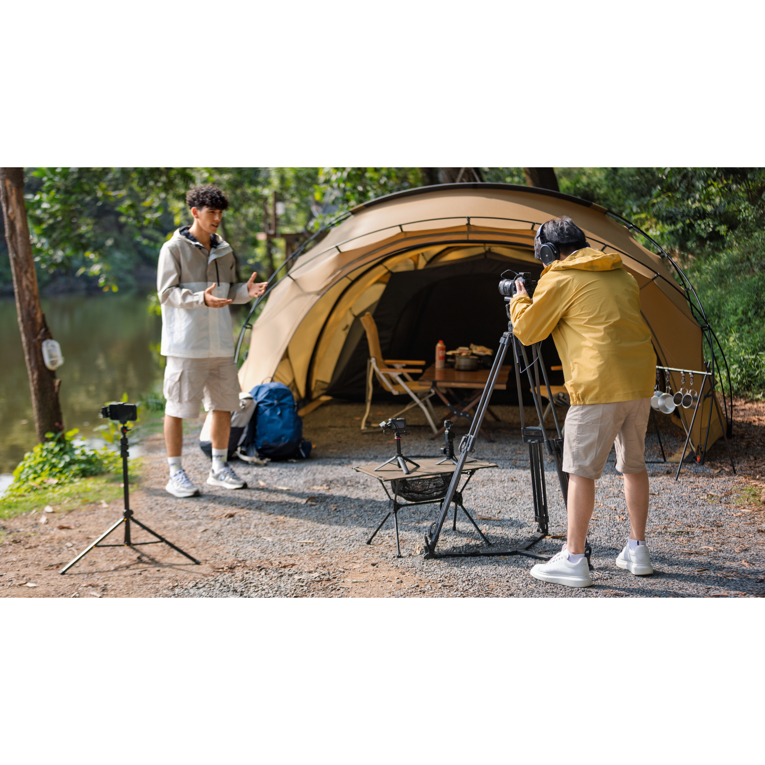 Two men filming in a campsite. One speaks, the other films. A tent, table, and tripod are visible, with trees and a lake in the background.