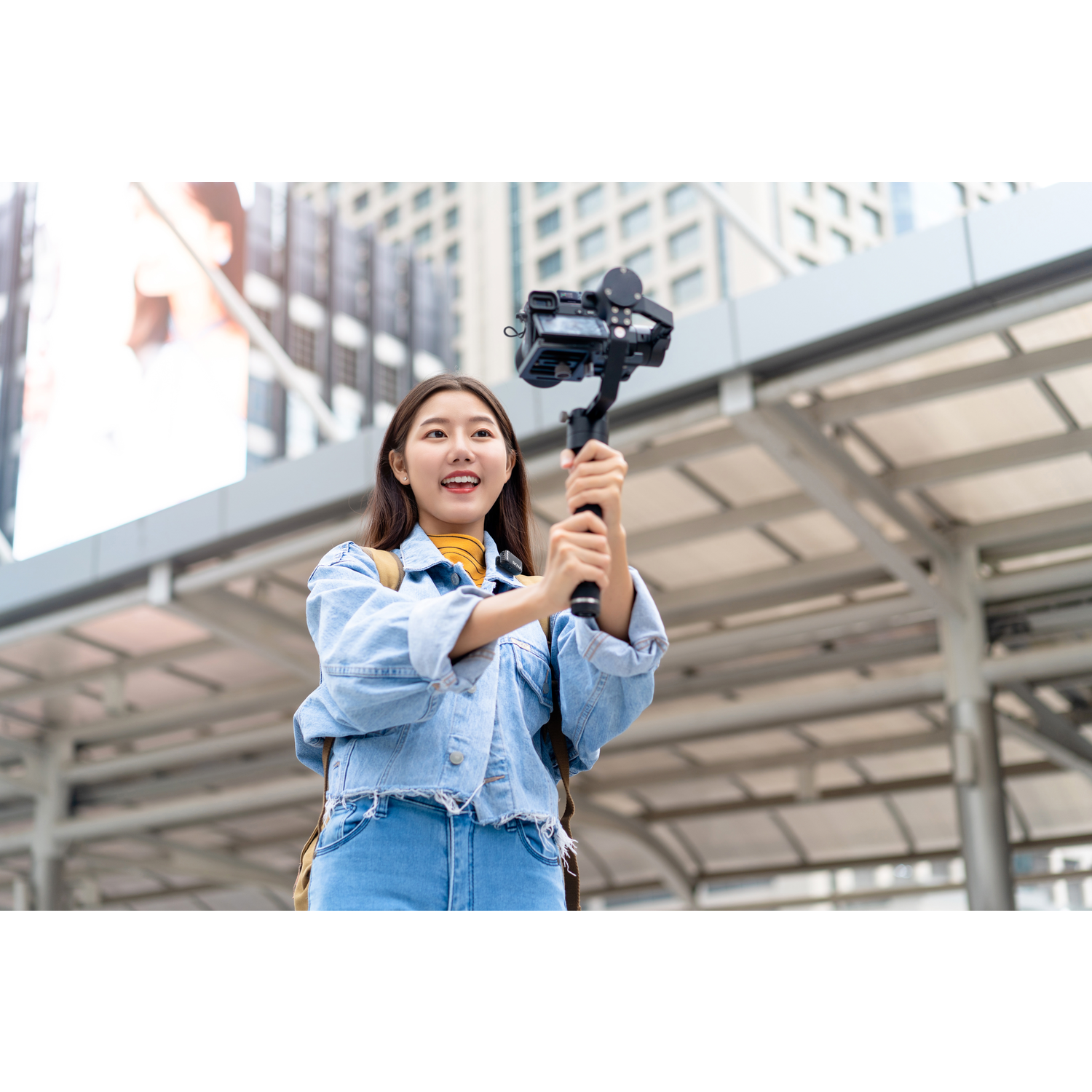 Young woman holds a camera on a gimbal, smiling. She wears denim and a yellow shirt. Background is urban with buildings.