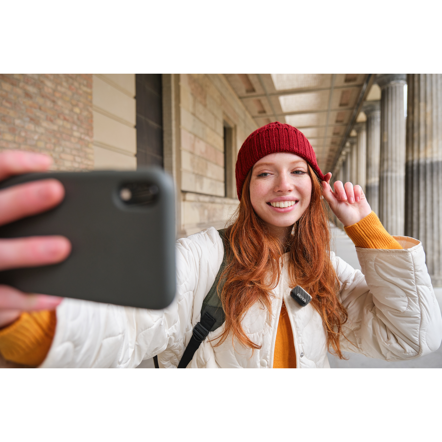 A smiling woman with red hair and a red hat takes a selfie. She wears a white jacket, yellow sweater, and stands outside a building.