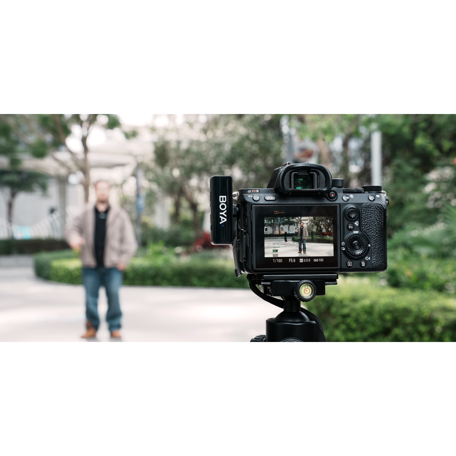 Camera on tripod filming a man in a park. Camera screen shows the man. Microphone attached. Green bushes and trees in the background.