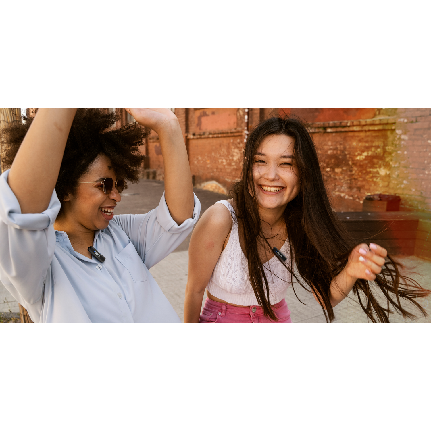Two young women laughing outdoors. One has afro hair and sunglasses, the other long hair and pink pants. They are wearing lavalier mics.
