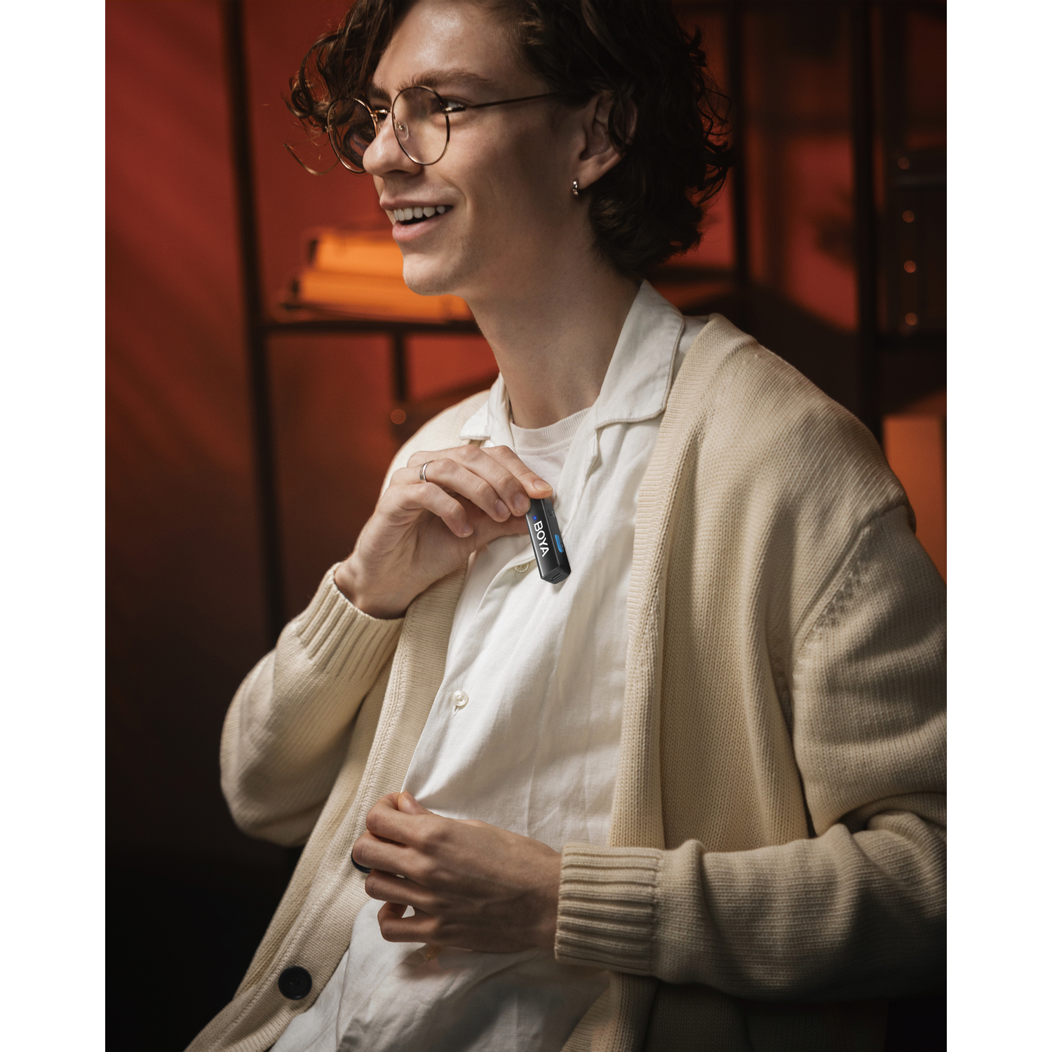 A man holding a BOYA microphone. He wears glasses, a white shirt, a cream cardigan, and smiles. The background is blurred and orange.