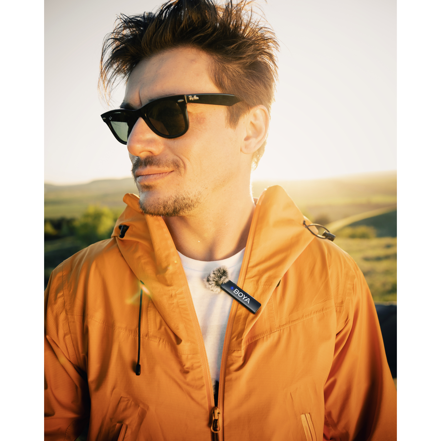 Man in sunglasses and orange jacket with a microphone attached. Background has green hills and bright sunlight.