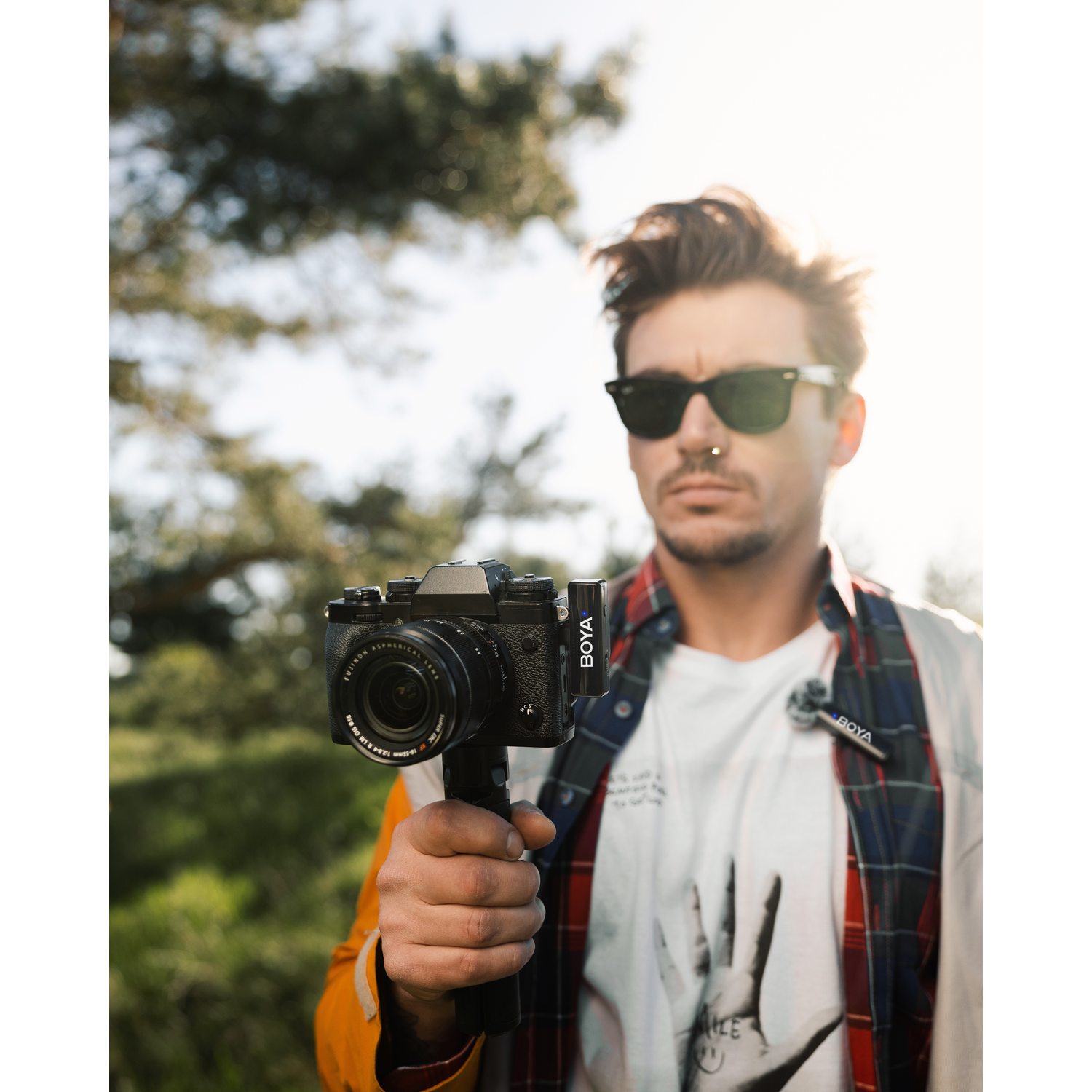 A man holds a black camera, wearing sunglasses and a plaid shirt. The background has trees and sunlight.