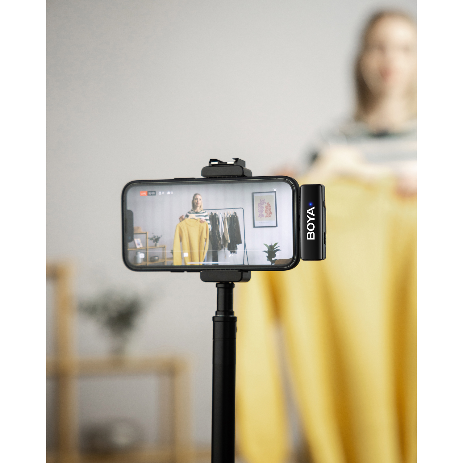 A woman is filming with a smartphone. The phone screen shows a yellow sweater. A Boya microphone is attached to the phone.