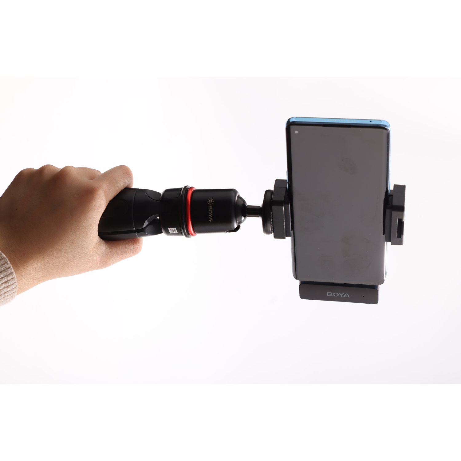 A hand holds a black phone holder with a smartphone. The holder has a red ring and the phone's screen is visible.