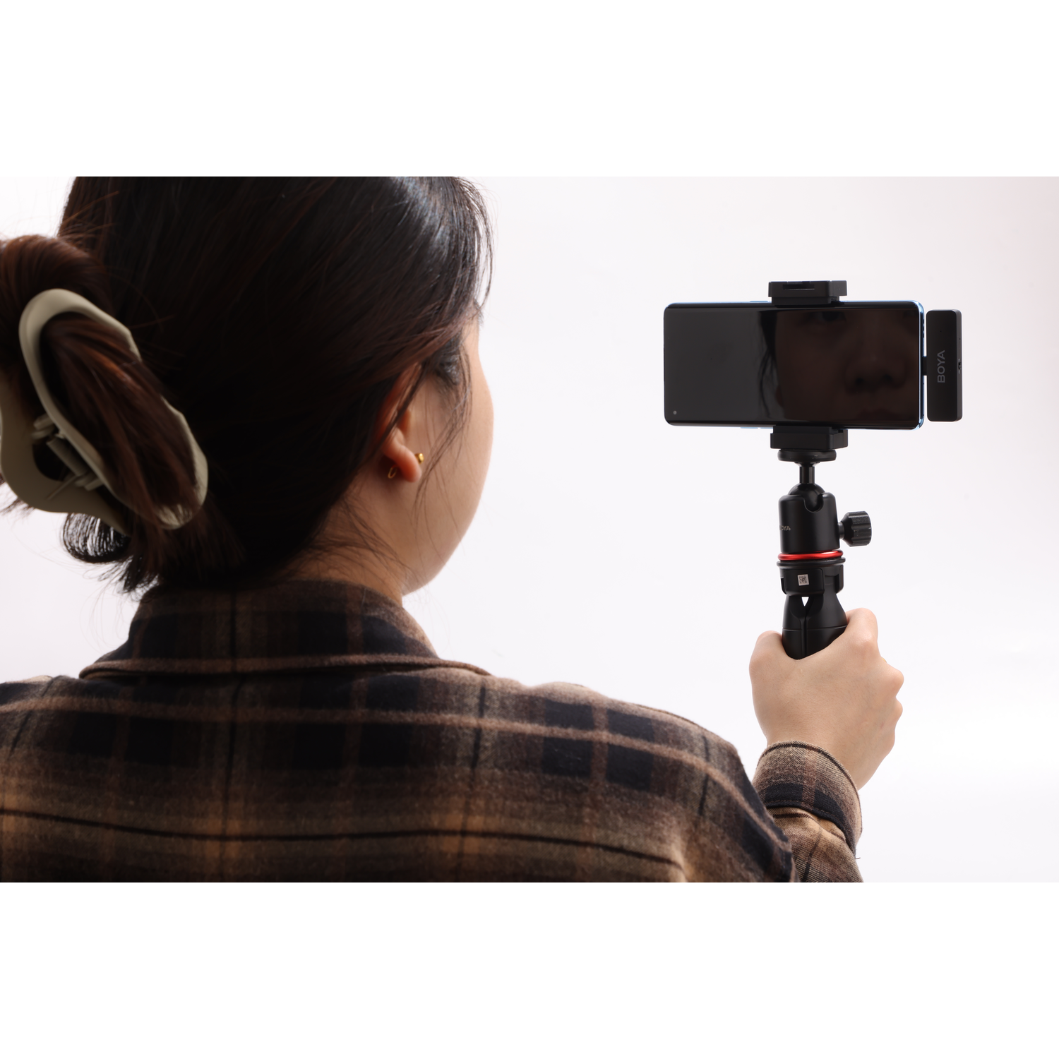 A person holds a phone on a tripod. The phone has a microphone attached. The person wears a plaid shirt, and hair is up.