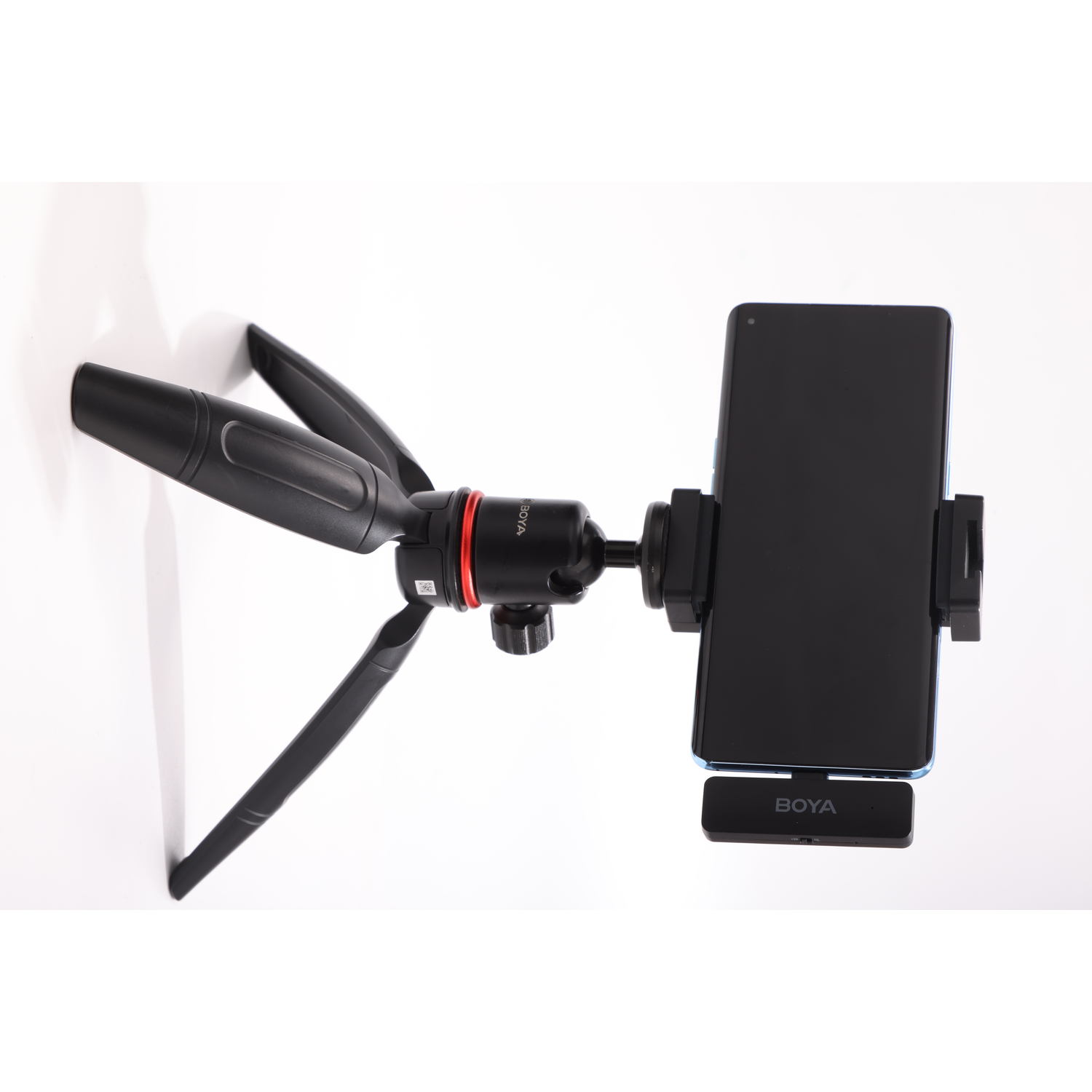 A black tripod holds a phone. A BOYA microphone is below the phone. The background is white.