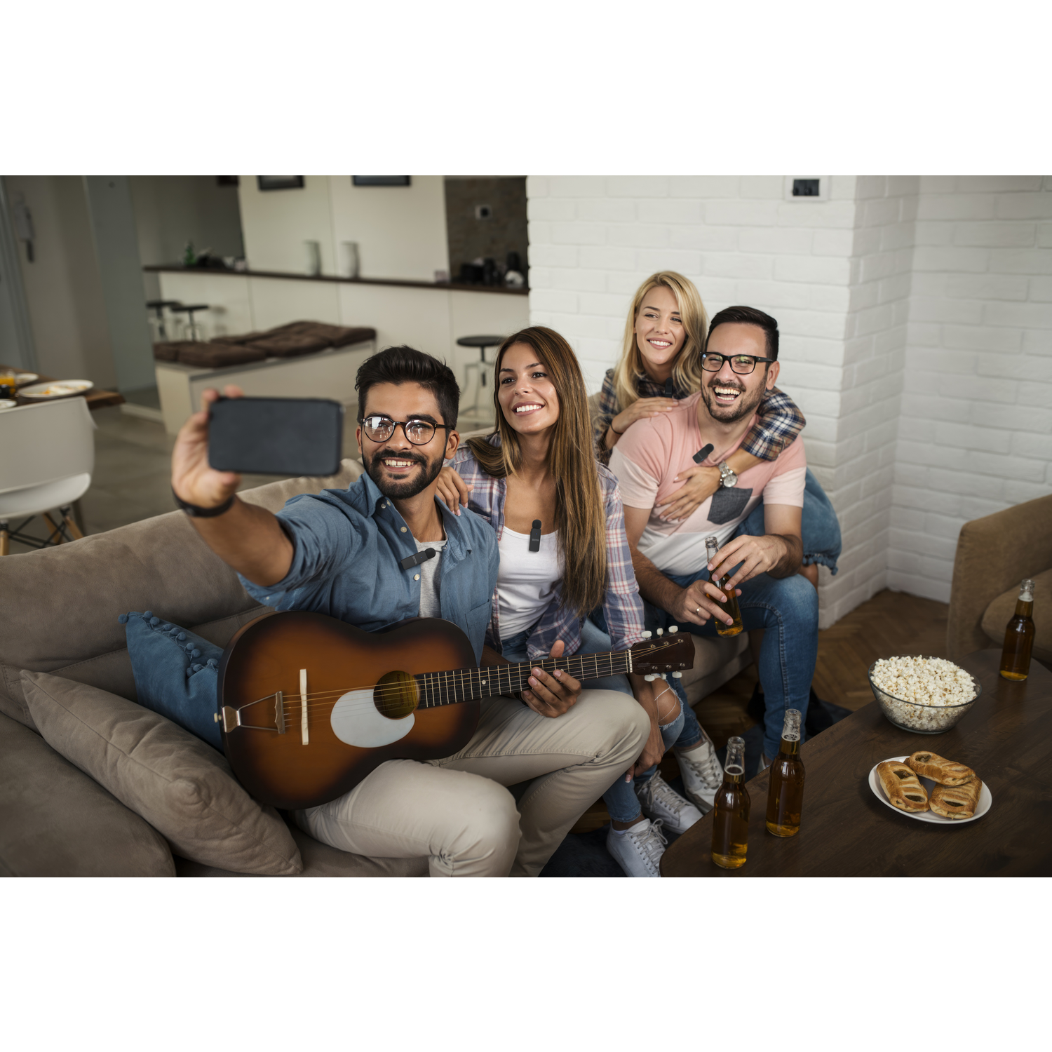 Four friends take a selfie. One holds a guitar. Beers, popcorn, and snacks are on a table. A cozy living room setting.