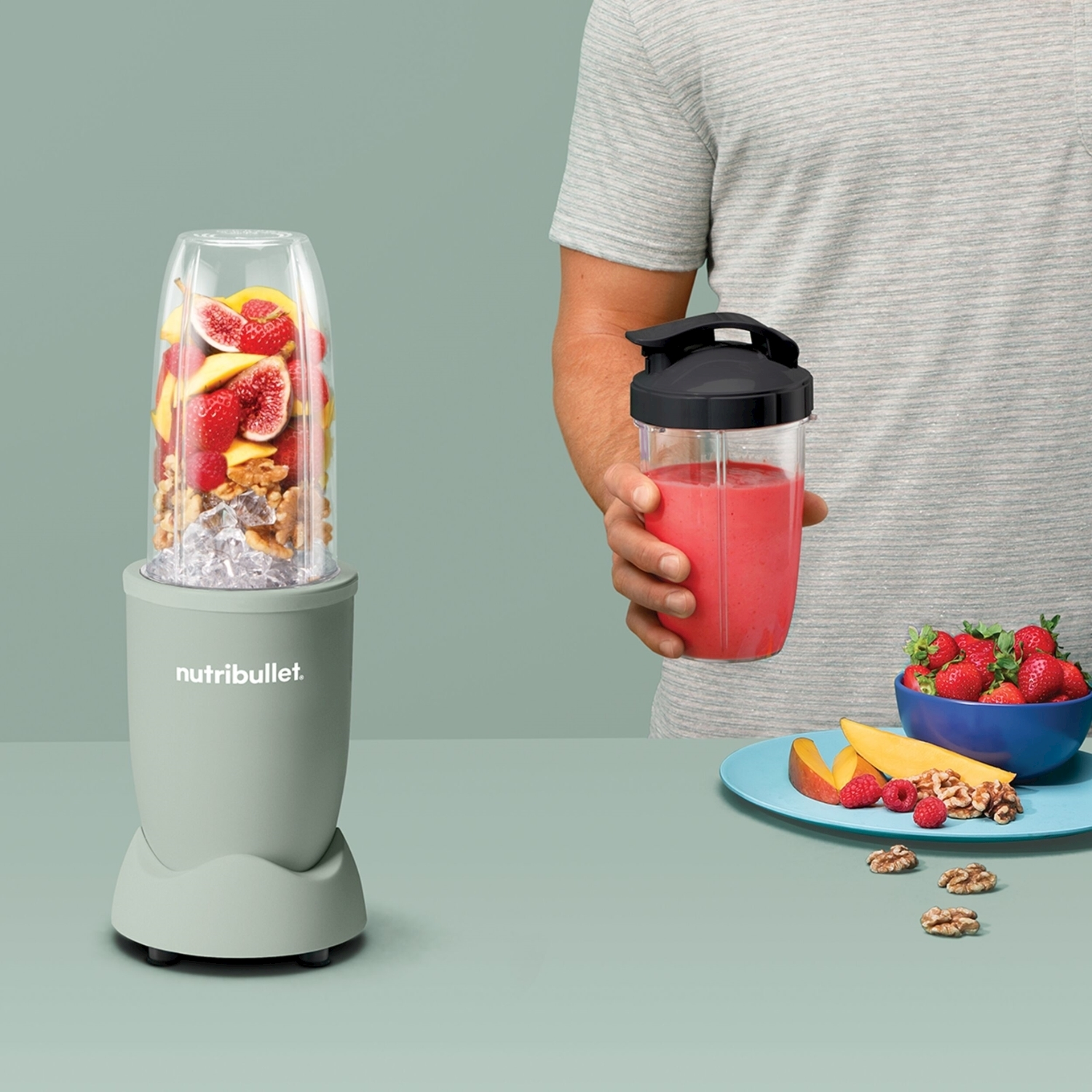 Una persona tiene in mano un frullato. Un frullatore con frutta, una ciotola con fragole e noci sono sul tavolo.