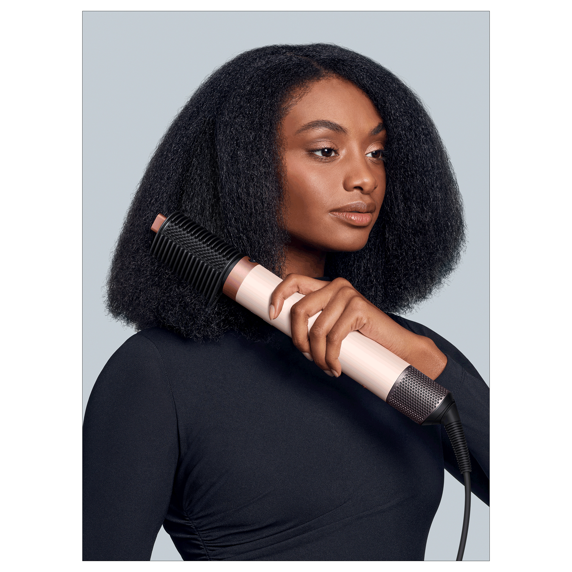 Kobieta z ciemnymi włosami trzyma narzędzie do stylizacji. Ma na sobie czarny top. Tło jest jasnoszare.