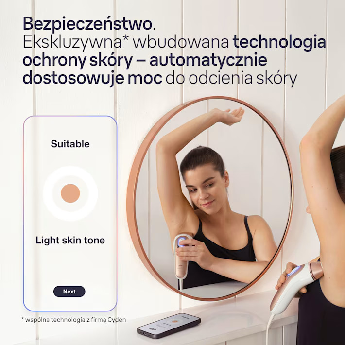 Kobieta używa urządzenia do usuwania włosów, odbitego w lustrze. Urządzenie jest pod jej ramieniem. Telefon leży na blacie.