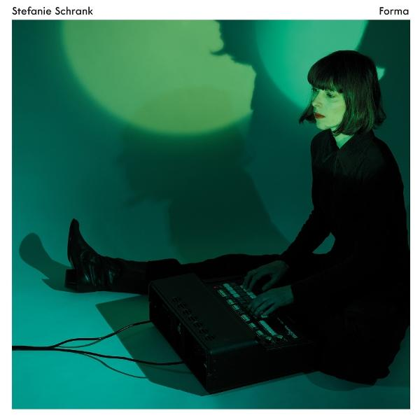 Eine Frau in Schwarz sitzt und spielt ein Synth-Keyboard vor einem grünen Hintergrund. Ihr Schatten wird an die Wand geworfen. Die Szene hat einen kühlen Ton.