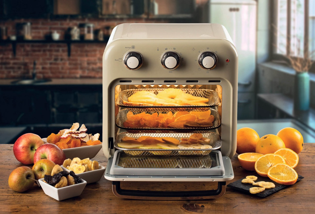 Frytkownica powietrzna z tacami jedzenia, otoczona owocami i suszonymi produktami. Tło przedstawia kuchnię z ceglaną ścianą.