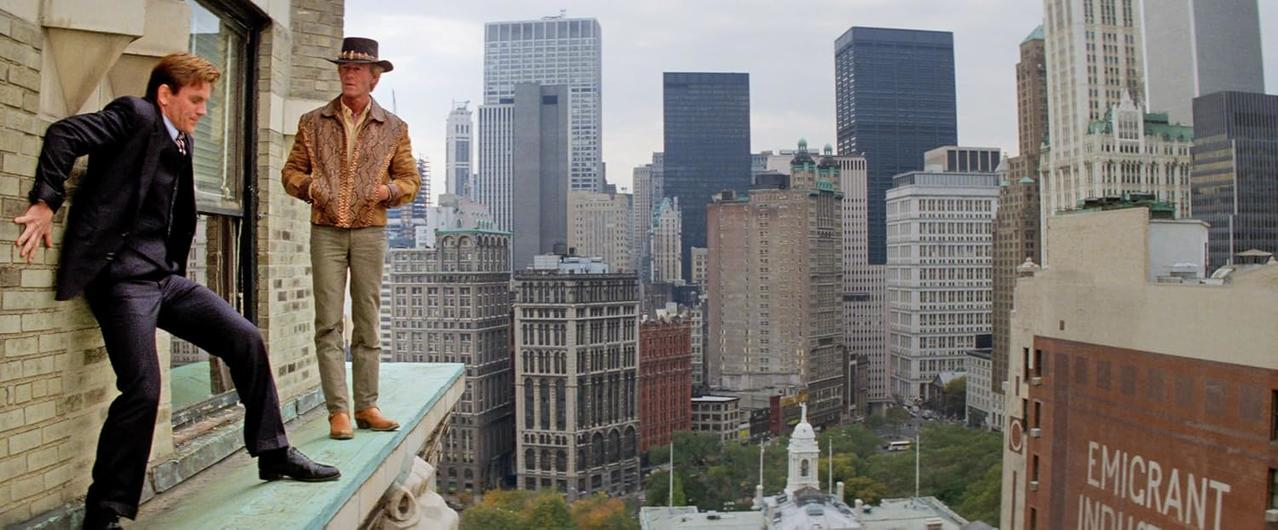 Zwei Männer auf einem Gebäudevorsprung in New York City. Einer klammert sich an die Wand, der andere trägt Hut und Jacke, dahinter die Skyline.