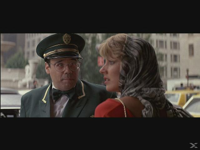 Ein Mann in grüner Uniform und eine Frau mit Schal. Sie sind draußen, mit Gebäuden und Taxis im Hintergrund.