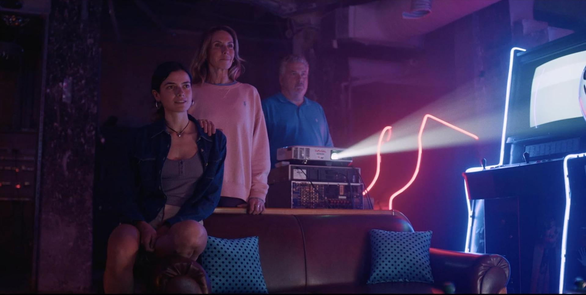 Drei Personen stehen in der Nähe eines Projektors und eines Retro-Spiels. Neonlichter und ein Ledersofa sind in der Szene zu sehen.