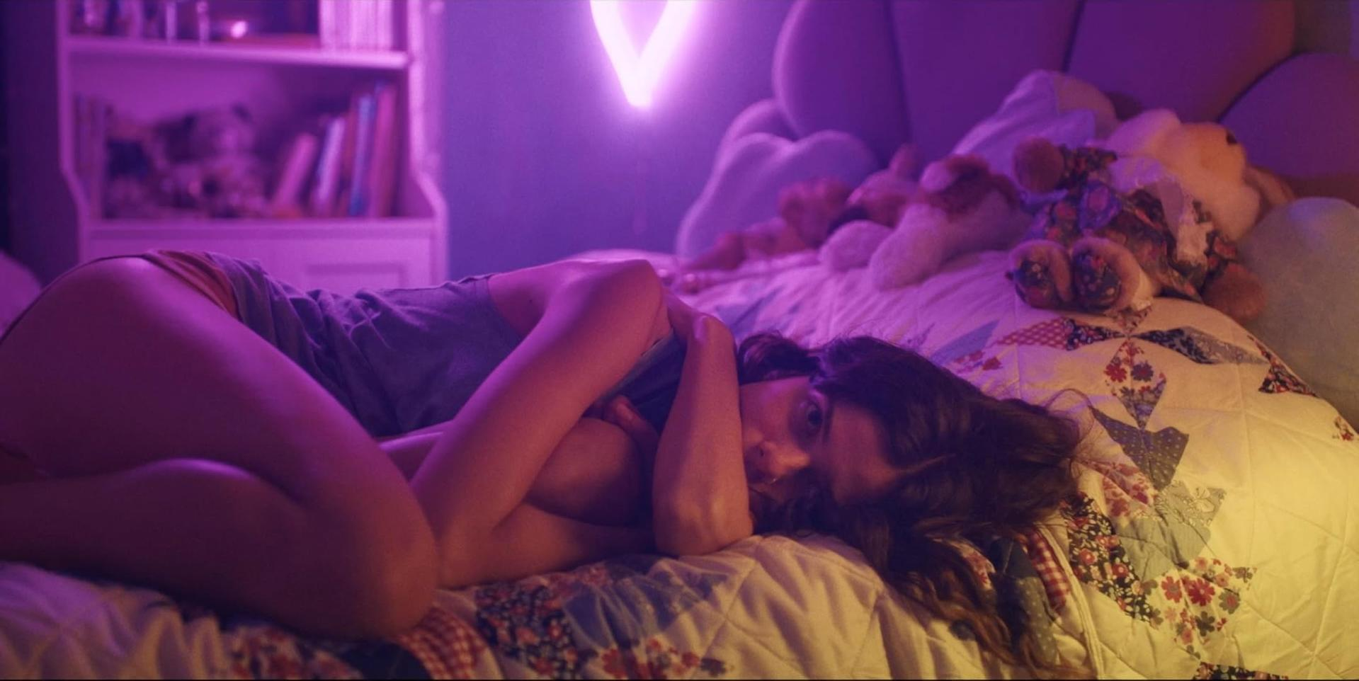 Eine Frau liegt auf einem Bett, die Arme verschränkt. Ein Neon-V ist an der Wand. Teddybären und eine gemusterte Steppdecke sind sichtbar.