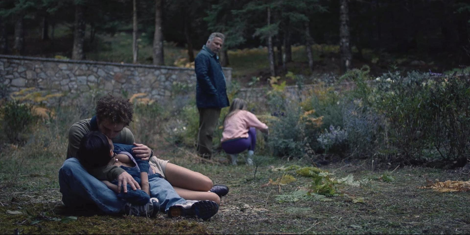Ein Mann sitzt und hält ein Kind in einem Wald. Ein anderer Mann und eine Frau sind im Hintergrund. Die Szene ist im Freien und natürlich.