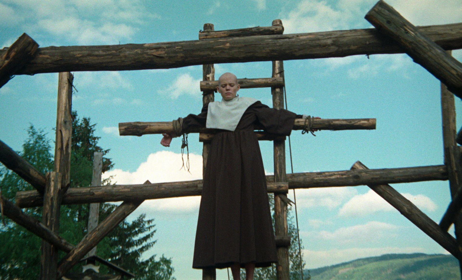Eine glatzköpfige Frau in einem braunen Kleid und weißem Kragen ist an ein Holzkreuz gebunden. Der Hintergrund zeigt einen Himmel mit Wolken und Bergen.
