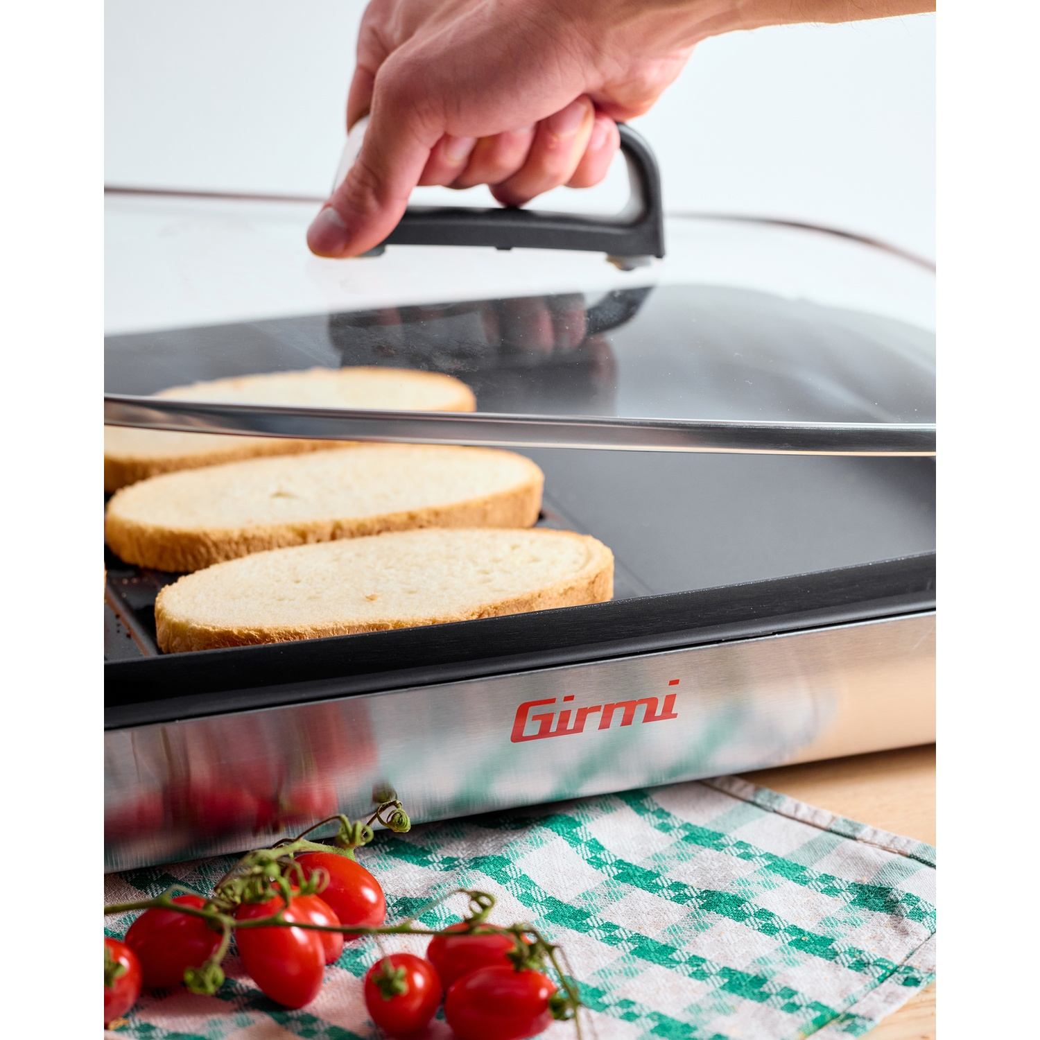 Mano che alza il coperchio di una piastra con fette di pane all'interno. Pomodori e un panno a scacchi in primo piano.