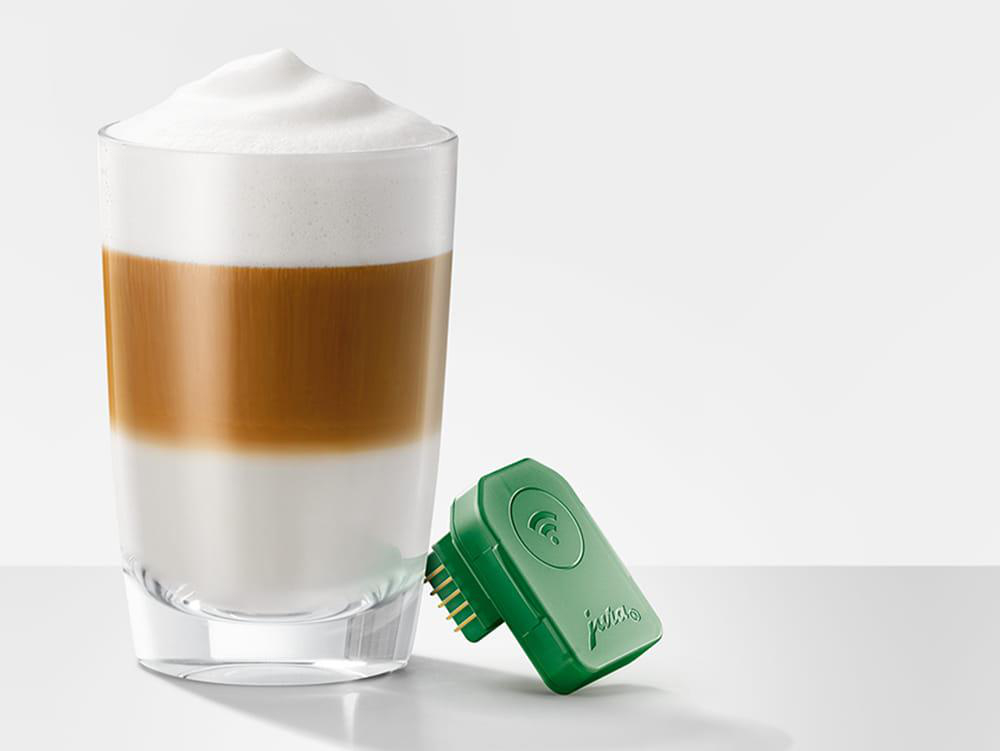 Ein Latte Macchiato-Glas mit Milch- und Kaffeeschichten, gekrönt von Schaum. Daneben liegt ein grünes Gerät mit einem WLAN-Symbol.