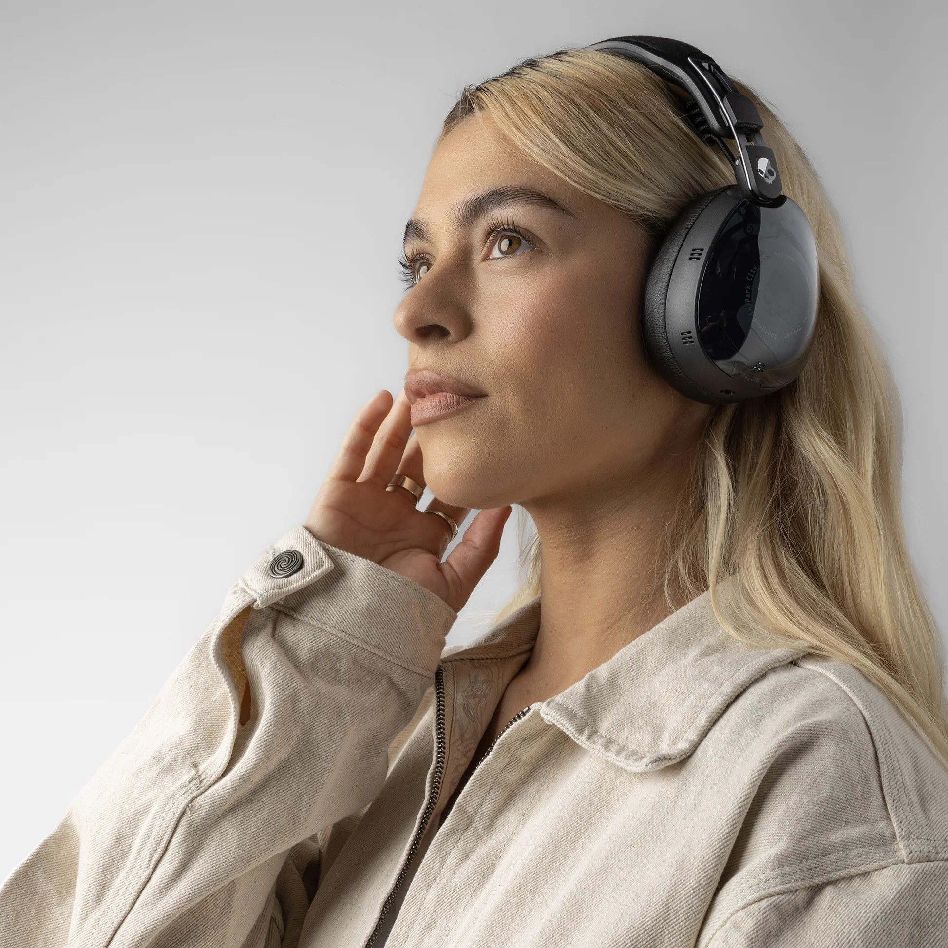 Blonde Frau mit schwarzen Kopfhörern, berührt ihr Gesicht. Sie trägt eine beige Jacke und blickt nach oben, mit weißem Hintergrund.