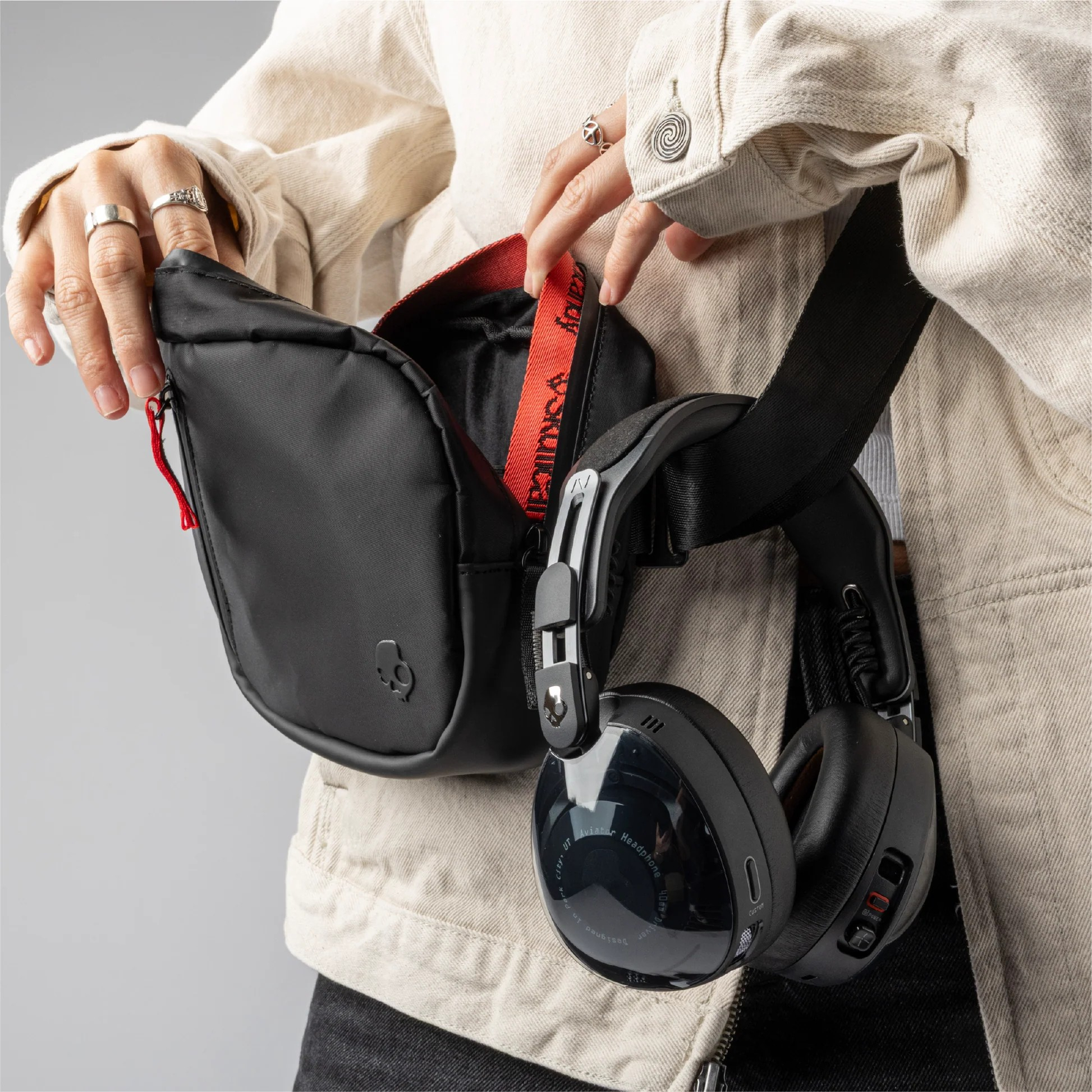 Person in beiger Jacke öffnet eine schwarze Bauchtasche. Kopfhörer hängen am Riemen. Ringe an den Fingern. Roter Riemen in der Bauchtasche.