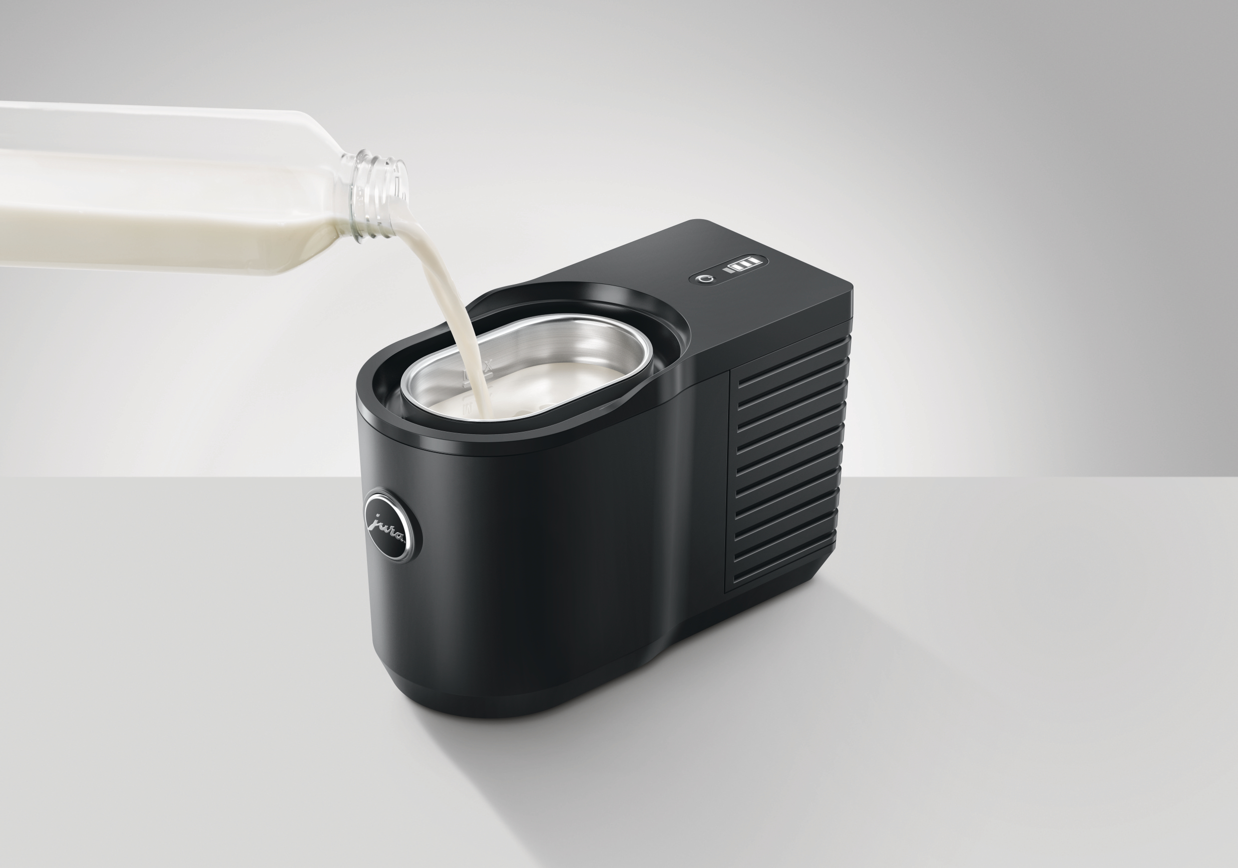 Ein schwarzer Milchaufschäumer wird mit Milch aus einer Glasflasche gefüllt. Die Maschine steht auf einer weißen Oberfläche.
