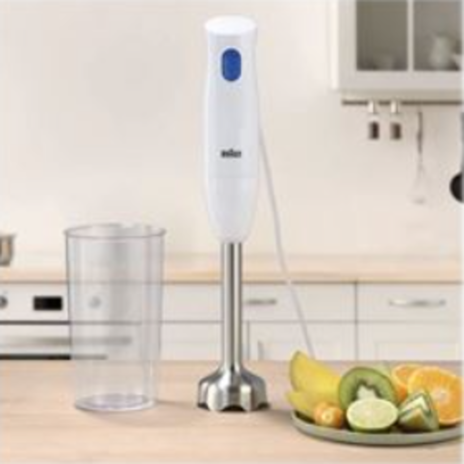 A white and silver hand blender stands on a wooden surface. A glass, and a plate of fruit are also present. Kitchen in the background.