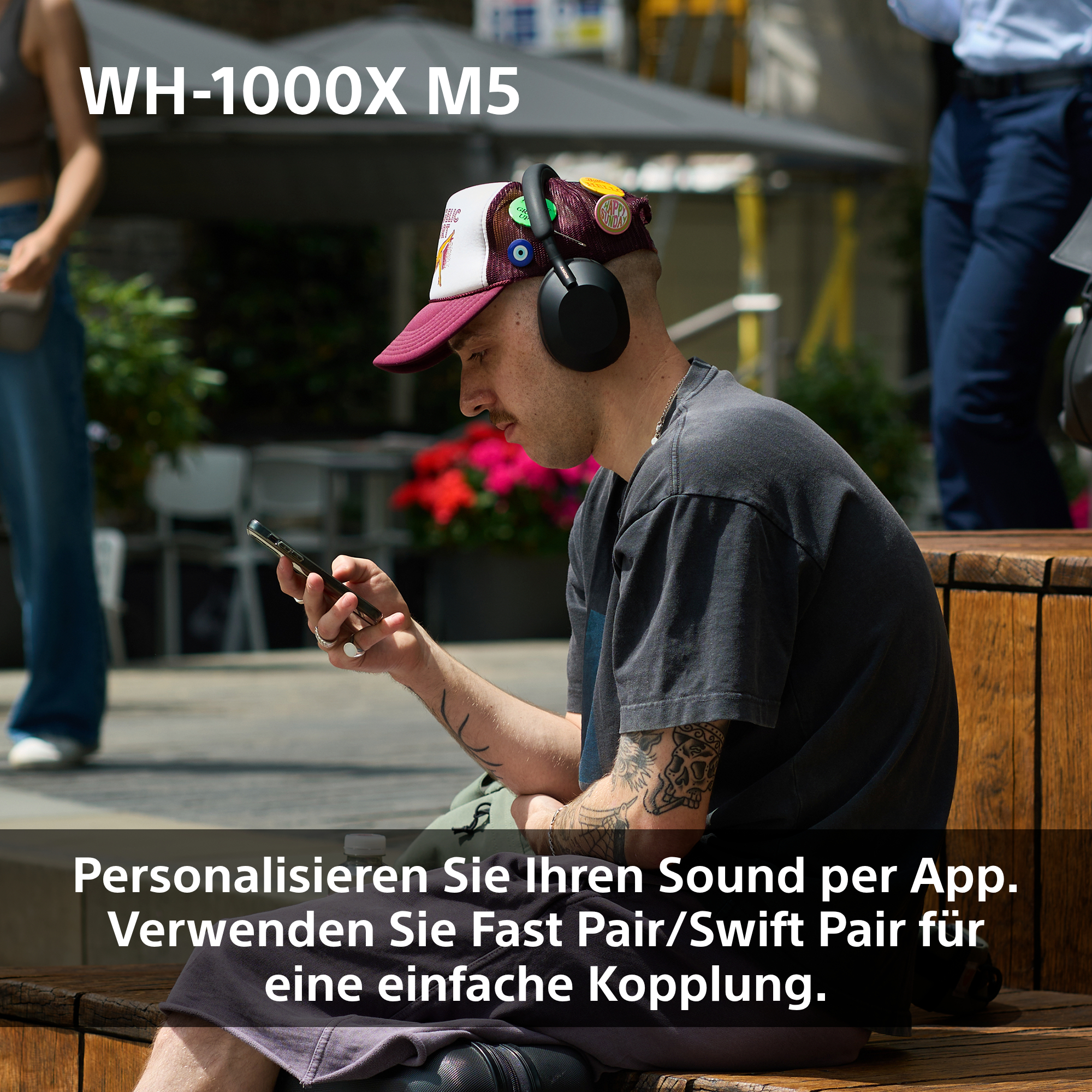 Mann mit Kopfhörern und Mütze, hält ein Telefon. Tattoos sichtbar. Im Hintergrund eine Straßenszene mit Menschen.