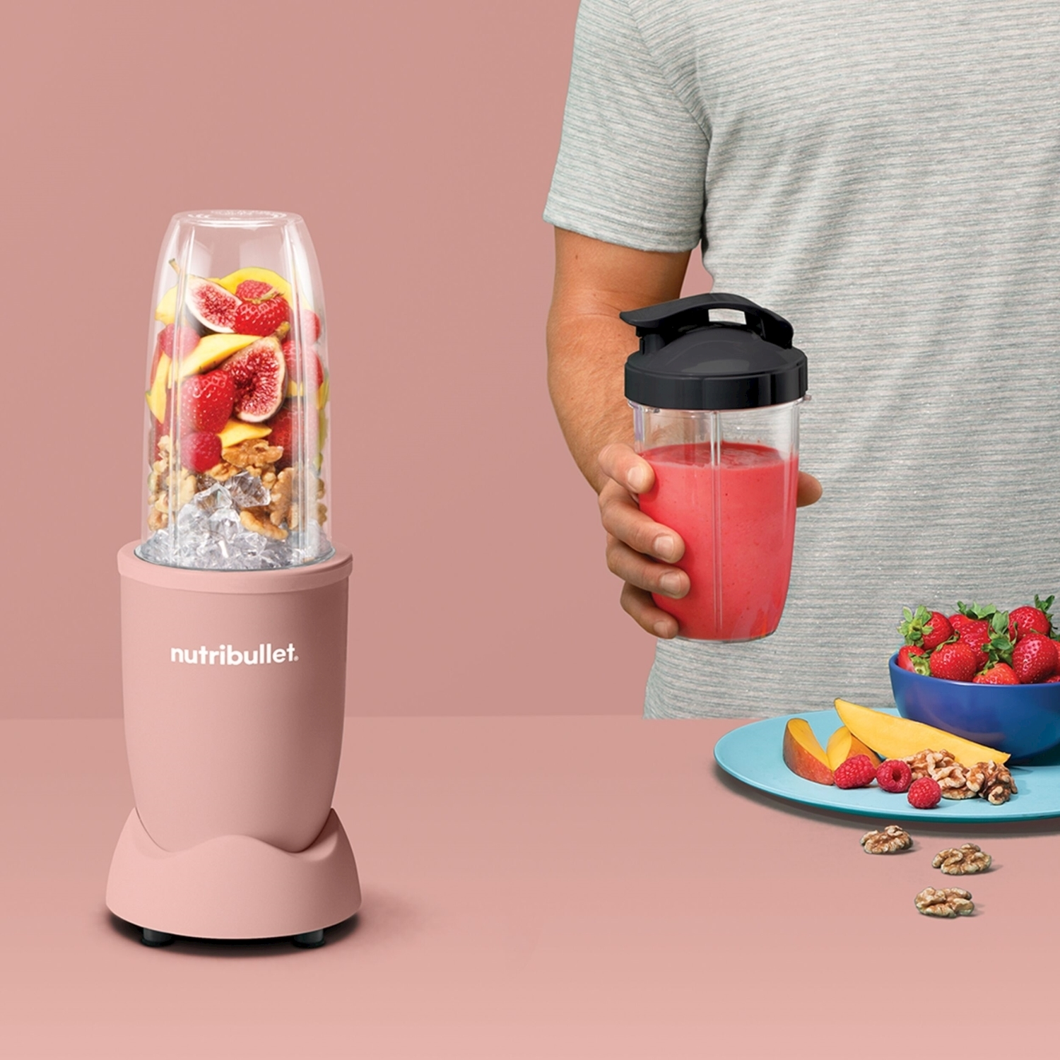 Un frullatore con frutta all'interno e una persona che tiene un frullato. Ci sono frutta, noci e una ciotola con fragole sul tavolo.