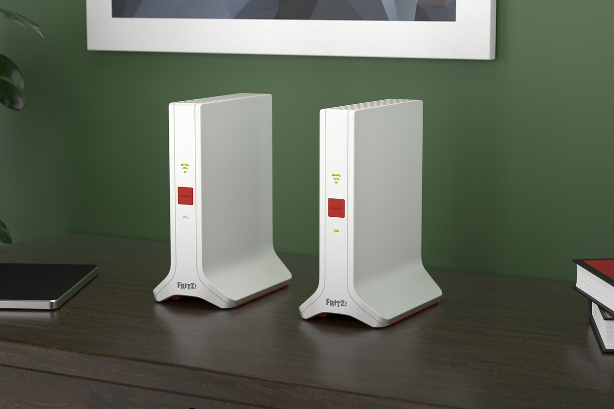 Due router bianchi con accenti rossi su un tavolo di legno scuro. Parete verde sullo sfondo con un quadro. Libri a destra.