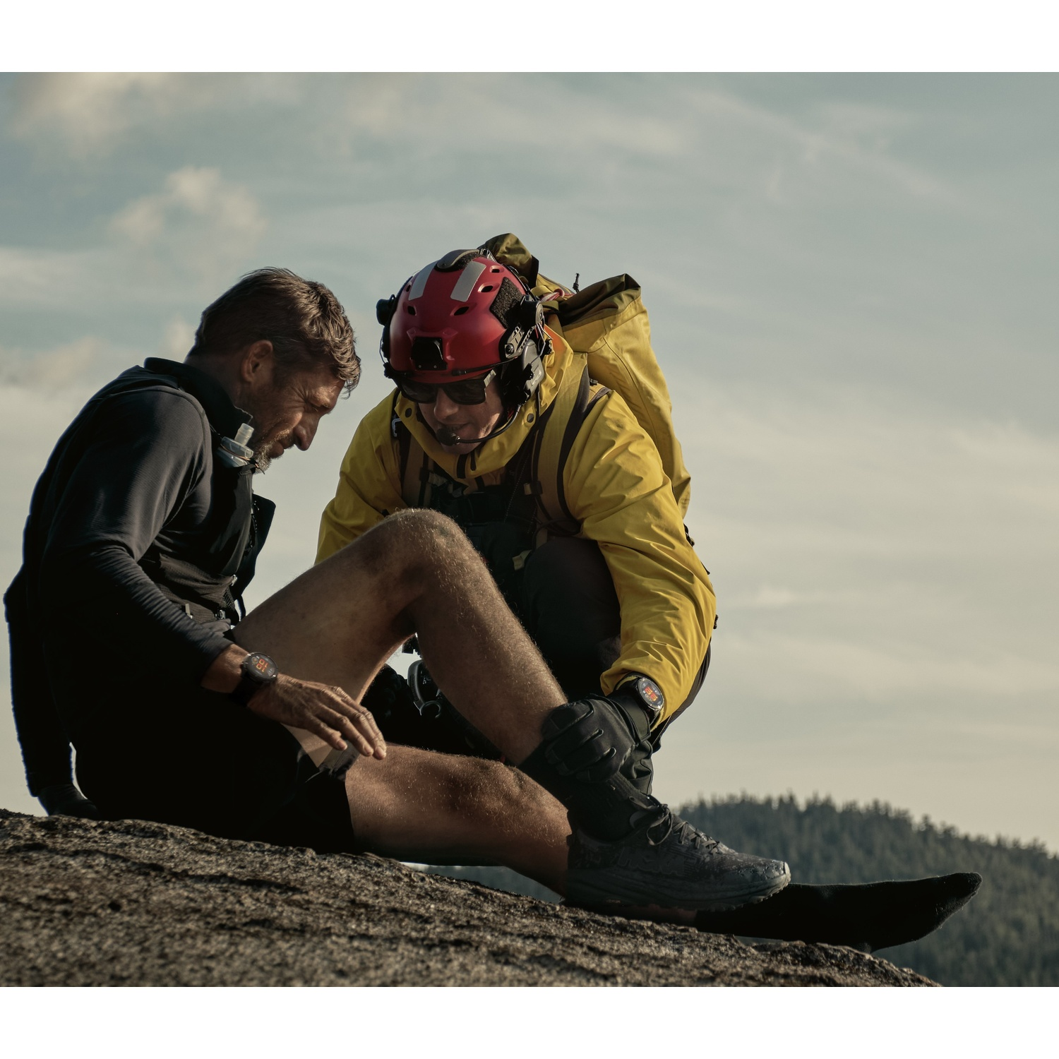 Deux personnes sur un rocher, une blessée. Une avec un casque rouge aide la personne blessée. Fond: forêt et ciel.