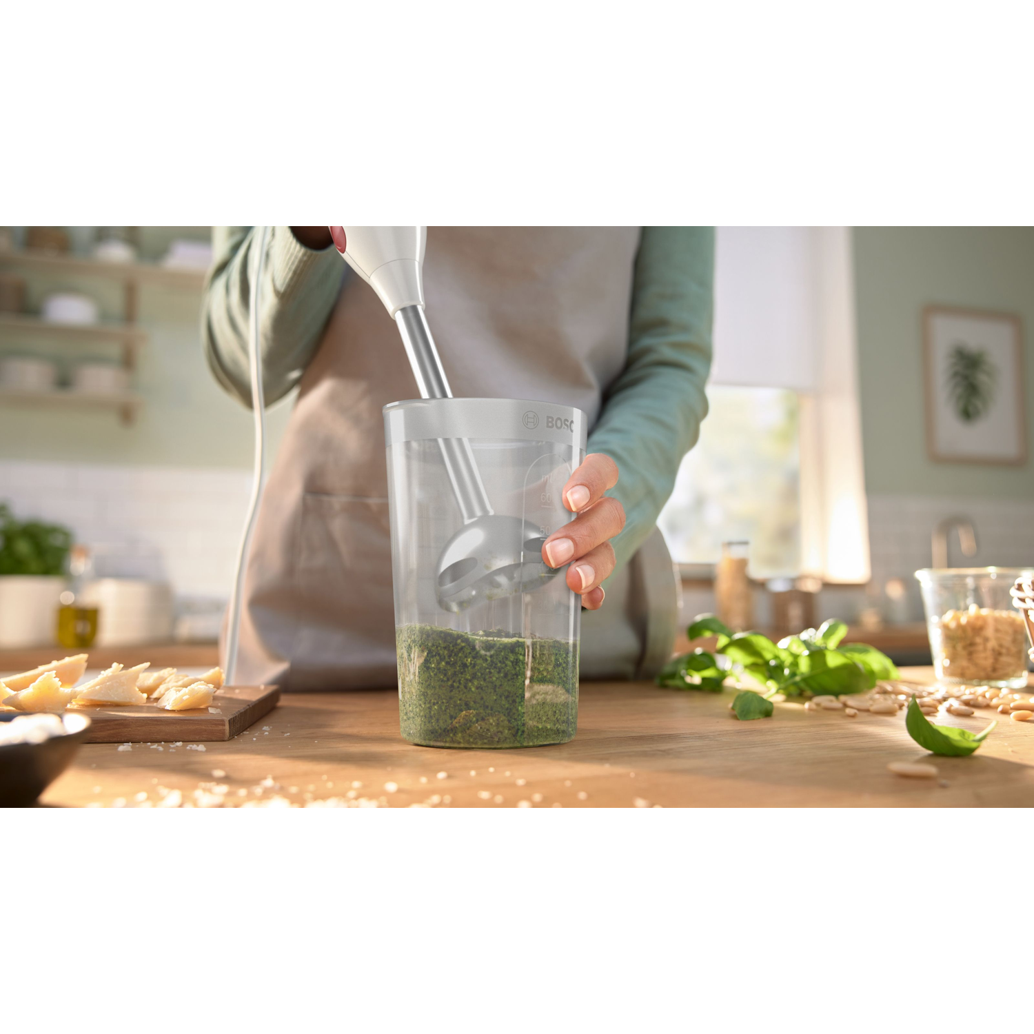Een persoon mixt pesto met een staafmixer. Ingrediënten zoals basilicum, pijnboompitten en Parmezaanse kaas liggen op de houten tafel.