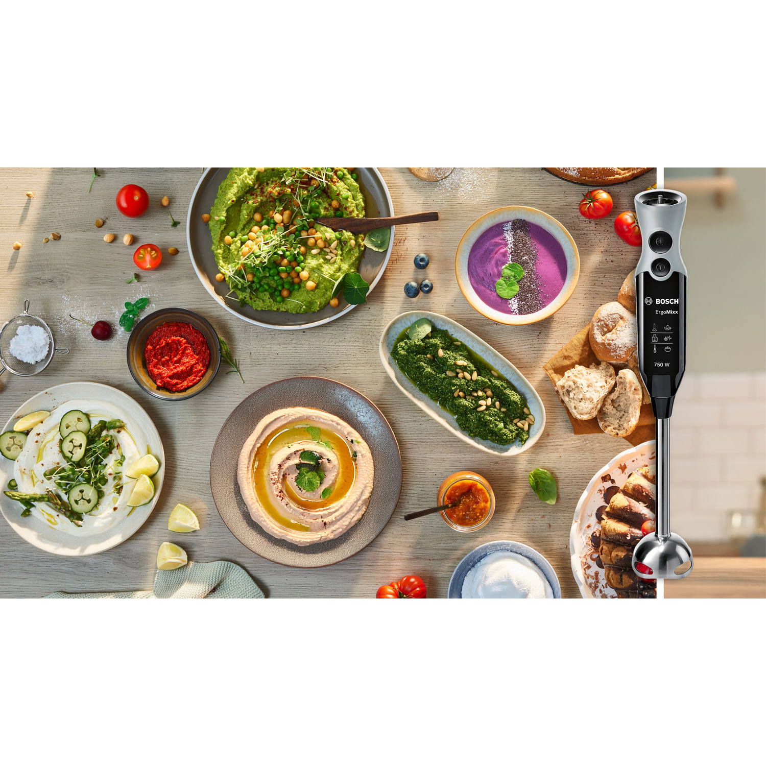 A wooden table is filled with various dishes including hummus, soup, pesto, and a Bosch ErgoMixx blender on the right.