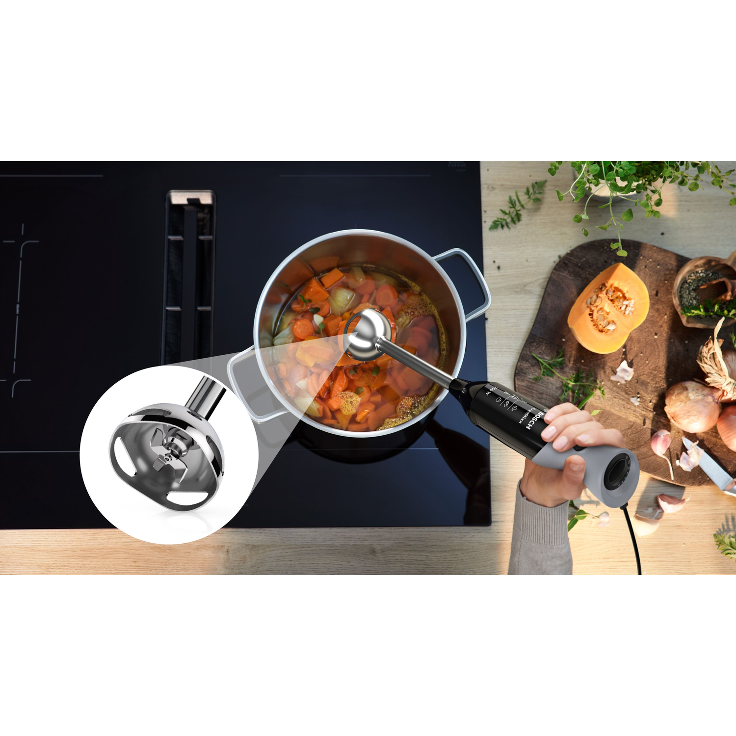 A hand holding a black immersion blender in use. A pot of soup simmers on a stovetop. Vegetables and herbs surround.