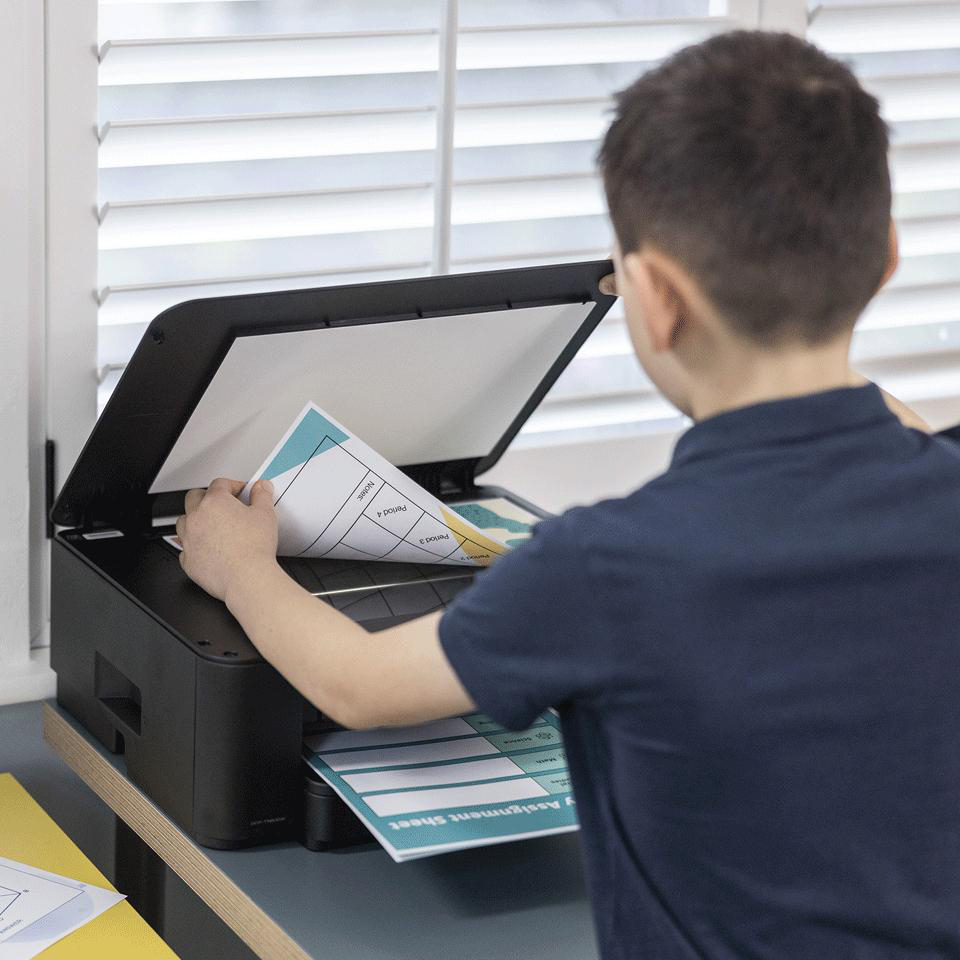 Ein Junge legt ein Arbeitsblatt in einen Drucker ein. Ein blaues Aufgabenblatt liegt im Drucker. Im Hintergrund sind Jalousien.