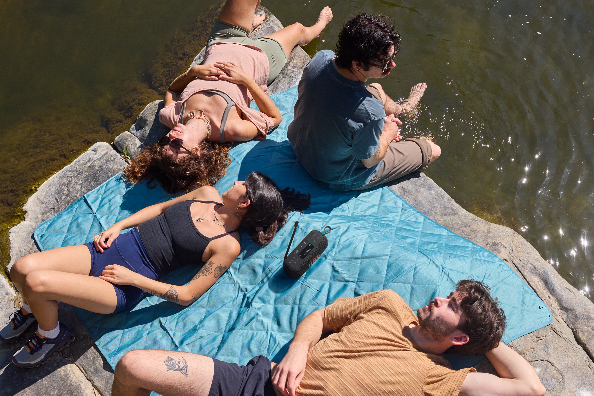 Quattro persone si rilassano vicino all'acqua in una giornata di sole. Una persona è seduta, altre sono sdraiate su rocce e una coperta blu. Un altoparlante è sulla coperta.