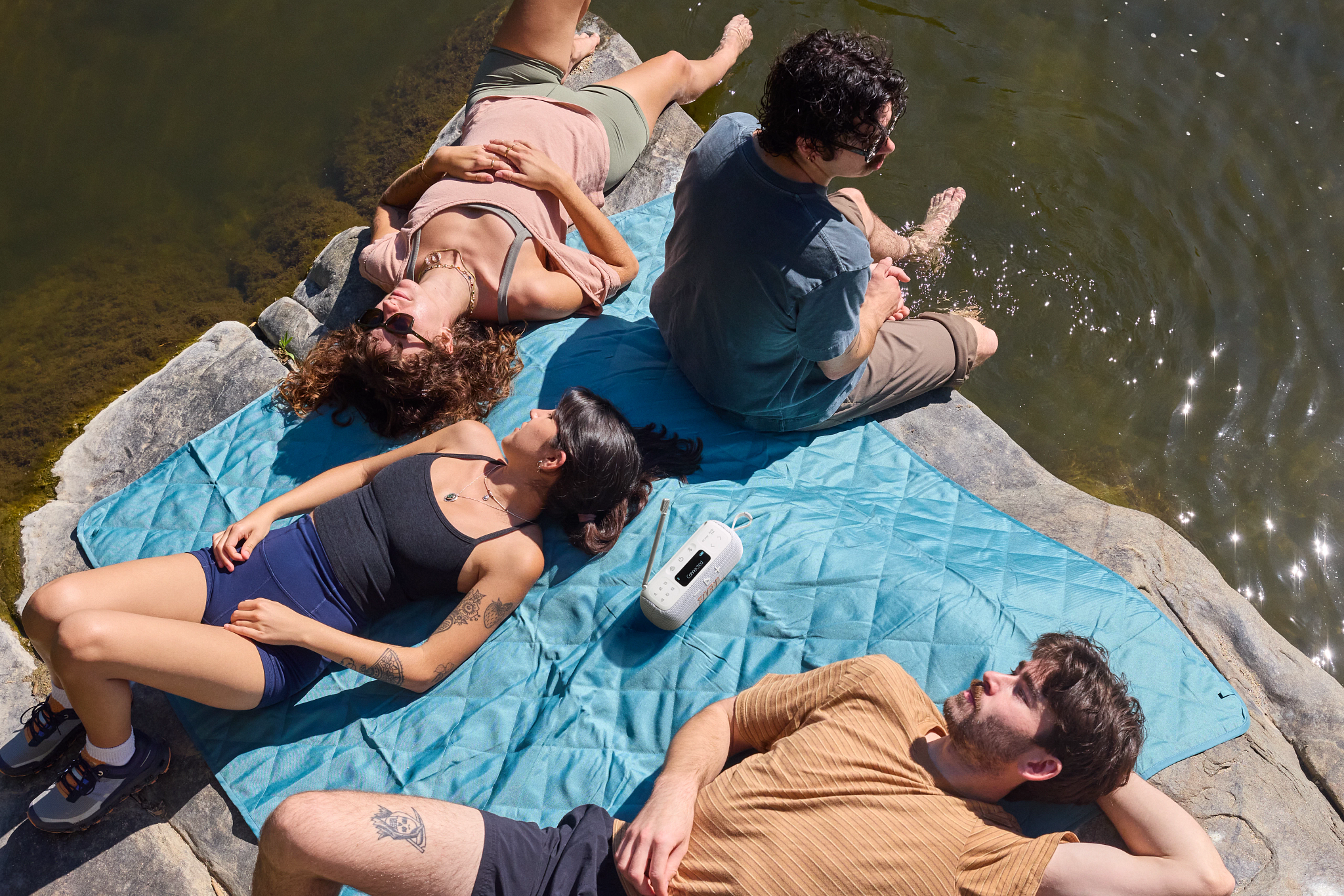 Quatre personnes se détendent au bord de l'eau, certaines sur une couverture bleue. Une personne est assise dans l'eau. Journée ensoleillée.