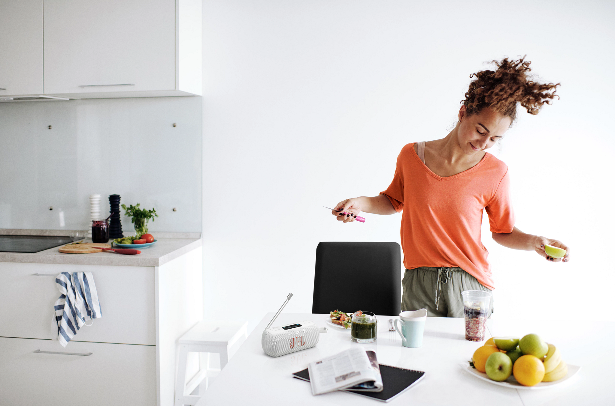 Une femme en chemise orange prépare de la nourriture dans une cuisine lumineuse. Sur une table, des fruits, une boisson et un haut-parleur JBL.