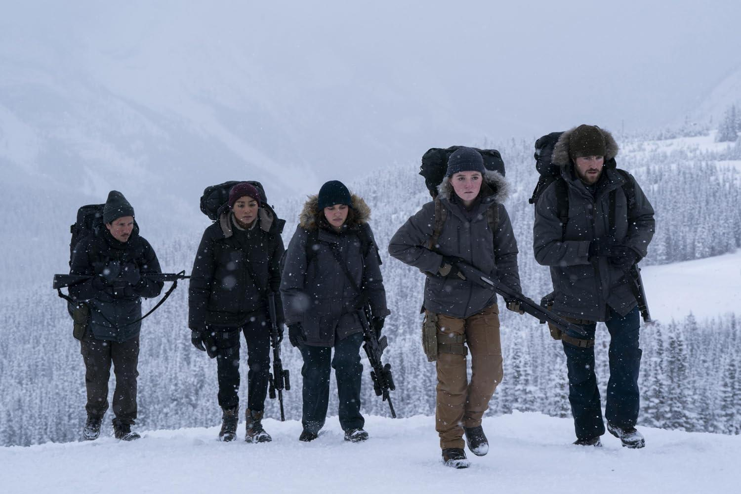 Fünf Personen in Winterkleidung gehen im Schnee. Sie tragen Rucksäcke und Waffen. Verschneite Berge im Hintergrund.
