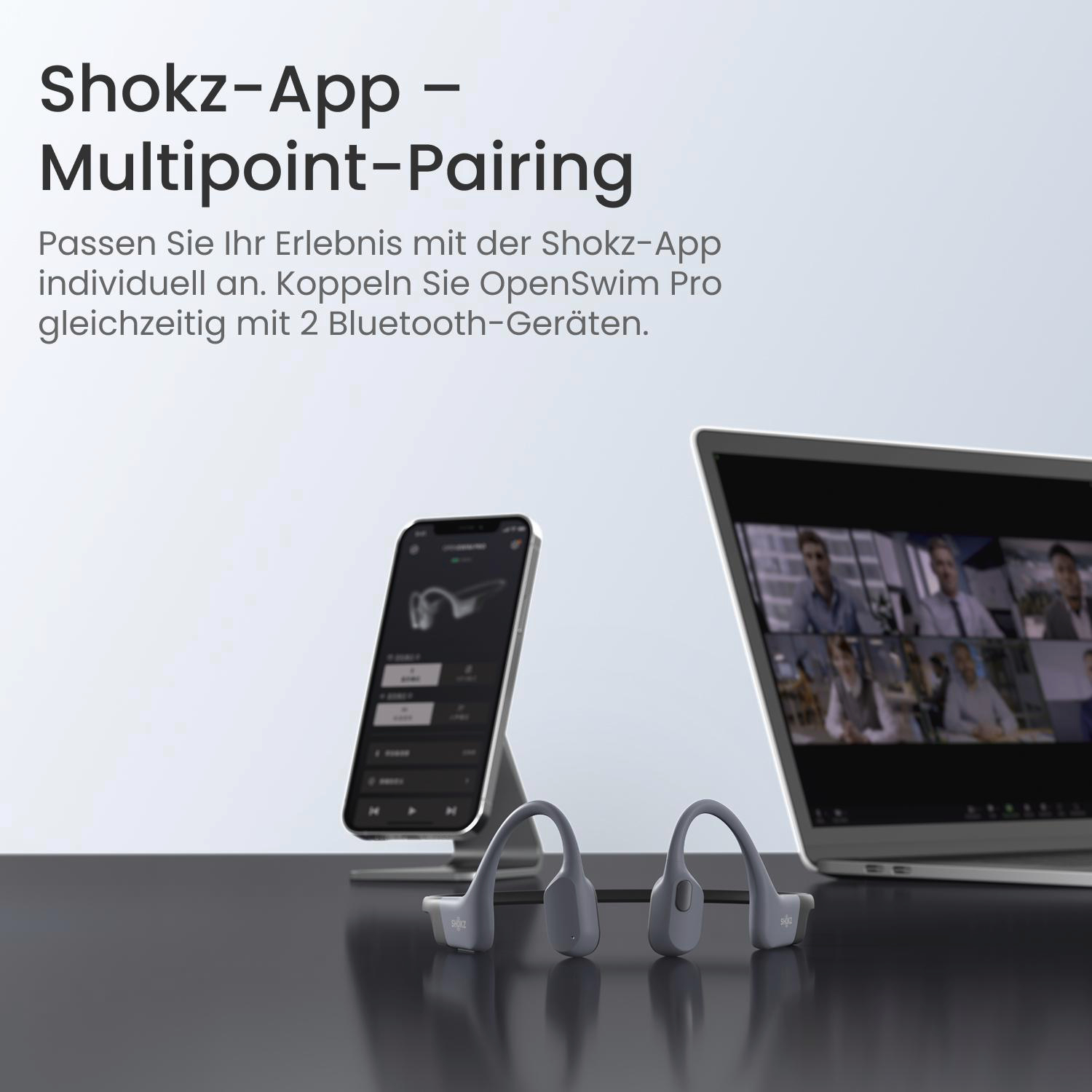 Ein graues Shokz-Headset und ein Smartphone auf einem Ständer stehen auf einer dunklen Oberfläche neben einem Laptop mit Videoanruf.