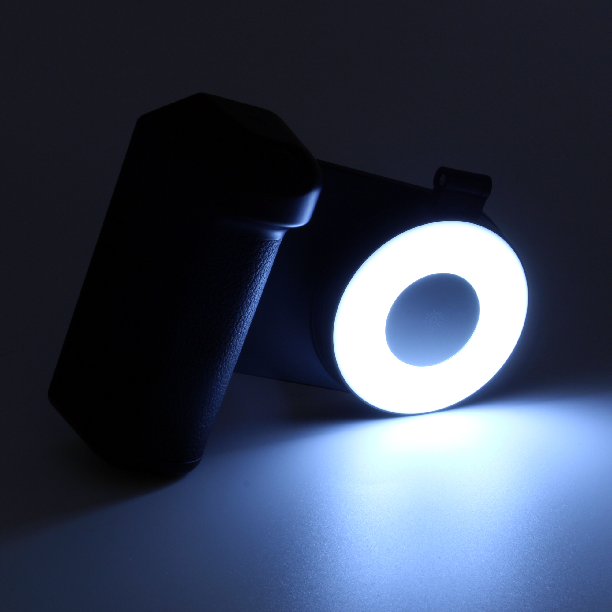 Ein schwarzes Ringlicht mit Griff liegt auf einer dunklen Oberfläche. Das Licht ist an und beleuchtet einen weißen Ring. Ein blauer Schein umgibt ihn.